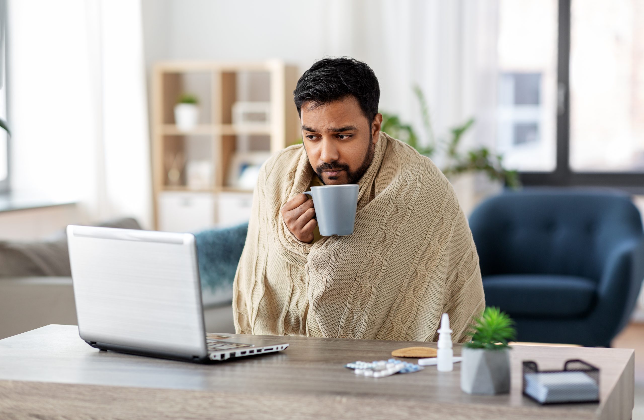 Man bundled up sick in a cold home with a laptop on the table in front of him.